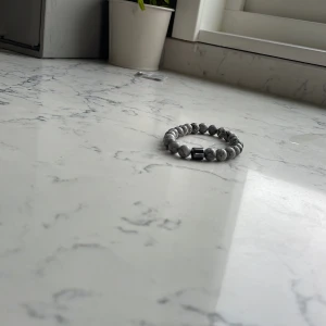 Gråmelerat pärlarmband med stenar - Snyggt armband med runda pärlor i gråmelerad stenlook och en svart cylinderformad detalj. Elastiskt band gör det enkelt att ta på och av. Perfekt accessoar för att lyfta din outfit med en cool och stilren vibe.