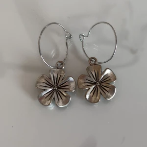 Silvriga örhängen med blomma - Ett par coola silvriga örhängen med stora blommor som hänger i tunna ringar. Blommorna har tydliga detaljer och ger en unik och trendig look. Perfekta för dig som vill sticka ut med ett smycke som känns både stilrent och lekfullt.