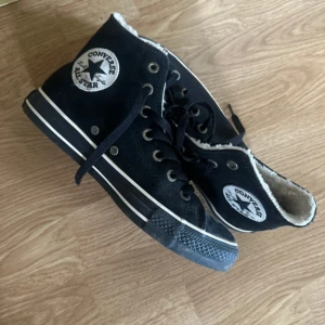 Svarta Converse All Star med foder - Svarta Converse All Star high-top sneakers med vitt teddyfoder på insidan. Klassisk rund tå, svart snörning och vit rand längs sulan. Perfekta för kyligare dagar när du vill ha både stil och värme. Ikonisk logga på sidan och slitstark gummisula.