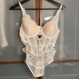 Vit spets body från Hunkemöller - Superfin vit korsett från Hunkemöller med vackra spetsdetaljer och transparent mesh. Den har bygel, reglerbara axelband och dekorativa sömmar som ger en snygg siluett. Perfekt för dig som vill ha något extra till outfiten.