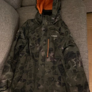 Camouflage vinterjacka från Everest - Säljer en grön och svart camouflage vinterjacka från Everest med orange foder. Jackan har huva, dragkedja med orange detalj och flera fickor. Perfekt för kalla dagar och har en cool streetstil.