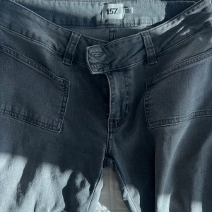 Mörkgrå jeansbyxor från 157 - Snygga mörkgrå jeansbyxor från 157 med raka ben och stora, fyrkantiga fickor framtill. Byxorna har dubbla knappar i midjan och klassiska bälteshällor. Materialet är mjukt jeans och färgen är diskret mörkgrå, perfekt för en chill och stilren look.