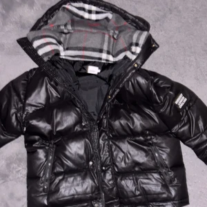 Svart pufferjacka från Burberry - Snygg svart pufferjacka från Burberry med huva och klassiskt rutigt foder. Jackan har dragkedja, stora fickor och en patch med Burberry London England på ärmen. Perfekt för kalla dagar och riktigt street i stilen.jätye billigt pris först till kvarn 