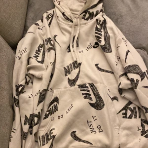 Nike hoodie med svart logga och print - Säljer en beige hoodie från Nike med svart, handritad logga och texten 'Just Do It' tryckt över hela tröjan. Tröjan har huva med snörning och en stor magficka. Perfekt för dig som gillar streetwear och vill sticka ut med en unik look.