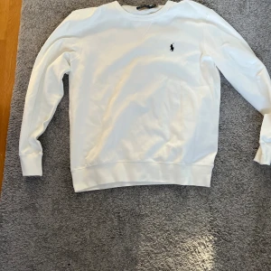 Vit sweatshirt från Ralph Lauren - Vit sweatshirt från Ralph Lauren med klassisk rund hals och broderad svart logga på bröstet. Tröjan har långa ärmar, ribbade muddar och är gjord i mjuk bomull. Enkel och stilren design som passar perfekt till jeans eller chinos.
