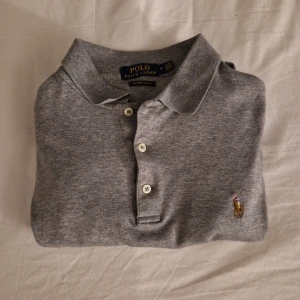 Grå långärmad pikétröja Polo Ralph Lauren - Snygg grå långärmad pikétröja från Polo Ralph Lauren i custom slim fit. Klassisk krage, tre knappar och broderad polospelare på bröstet. Tillverkad i mjuk bomull som känns skön mot huden. Perfekt för en clean och stilren look.