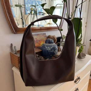 Mörkbrun axelväska - Väska från Nakd. Ny utan prislapp, aldrig använd. Brun. Justerbart handtag mellan 17-28,5 cm. Höjd: 15cm. Bredd 12cm. Längd: 38cm. Stort innerfack med två små fack, ett dragkedja. Silvriga detaljer. Inköpt för 399kr. 