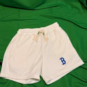 Vita MLB shorts med blå B-logga - Snygga vita shorts från MLB med blå B-logga broderad på vänster ben. De har elastisk midja med snörning och tofsar, samt fickor på sidorna och en bakficka. Perfekta för chill dagar och sportiga vibes.Liknande modell som originalet!