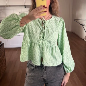 Grön- och vitrandig blus med snörning - Säljer en söt blus i ljusgrönt och vitt med tunna ränder. Blusen har voluminösa långa ärmar, snörning framtill och en lös flowy passform med en lätt croppad längd. Den har tyvärr inte kommit till användning💓