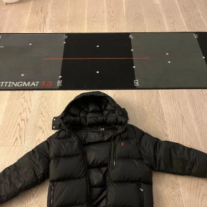 Svart pufferjacka från Polo Ralph Lauren - Svart pufferjacka från Polo Ralph Lauren med röd broderad logga på bröstet. Jackan har huva, dragkedja framtill och två sidofickor med dragkedja. Perfekt för kalla dagar och riktigt snygg streetstil. Jackan är väldigt sparsamt använd. Priset är inte hugget i sten, tveka inte på att höra av er!!