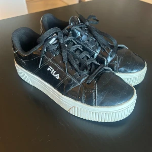 Svarta sneakers från Fila - Snygga svarta sneakers från Fila med croc-mönstrad yta och vita, tjocka sulor. Skorna har snörning och Fila-logga på sidan och bak. Perfekta för dig som vill ha en cool och trendig look med lite extra edge.