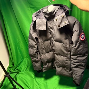 Grå jacka   Canada Goose - Säljer en grå dunjacka från Canada Goose med huva och snygga detaljer som tryckknappar och dragkedja vid fickorna. Jackan har ett klassiskt märke på ärmen och är perfekt för kalla dagar. Passformen är normal och jackan är riktigt varm och stilren.Liknande modell som originalet!