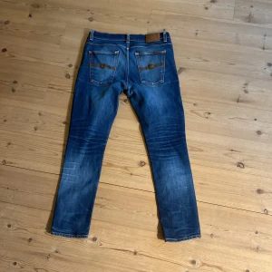 Blå jeans från Nudie, slim fit - Säljer ett par klassiska blå jeans från Nudie Jeans med perfekta  slitningar och . Modellen har smal passform, fem fickor och ikoniska broderade bakfickor. Jeansen är i bomull och har en mörkblå tvätt med coola detaljer. W34 men passar som 32  