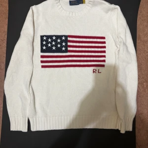 Ralph lauren stickat tröja - Vit stickad tröja från Polo Ralph Lauren med amerikansk flagga och RL-broderi på bröstet. Tröjan har rund halsringning, ribbade muddar och är tillverkad i bomull. Perfekt för dig som vill ha en klassisk och stilren look med en cool detalj framtill.