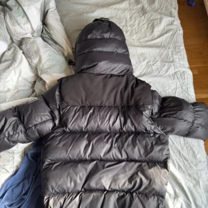 Svart Burberry pufferjacka med huva - Svart pufferjacka från Burberry med stor huva och klassiskt rutigt foder. Jackan har dragkedja och knappar framtill, två sidofickor med dragkedja och en patch med Burberry London England på ärmen. Perfekt för kalla dagar och riktigt snygg streetstil.                                                                 Pris går att diskutera vid snabb affär och hör av dig vid frågor😁