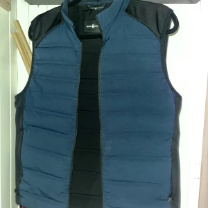 Blå dunväst från Sail Racing - Snygg blå dunväst från Sail Racing med svarta detaljer på axlar och sidor. Västen har quiltad design och dragkedja framtill, perfekt för lager-på-lager. Stilren look som funkar till många outfits. Märket syns tydligt på insidan. Skicket så gott som ny men den är använd. 