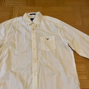 Vit skjorta från GANT i bomull - Klassisk vit skjorta från GANT med button down-krage och bröstficka med broderad logga. Skjortan är i bomull och har lång ärm samt regular fit. Perfekt för en clean och stilren look.