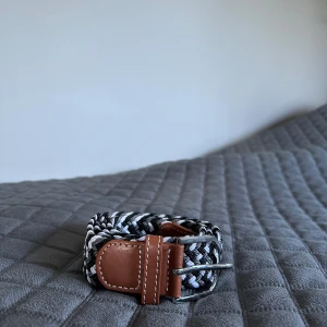 Svartvittgrått  flätat bälte med skinn - Snyggt flätat bälte i svart, vitt och grått med detaljer i brunt skinn och silverspänne. Bältet har ett coolt mönster och passar perfekt till jeans eller chinos. Skinnet har vita sömmar som ger en schysst kontrast mot det flätade bandet.