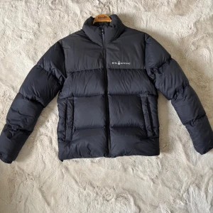 Svart pufferjacka från Sail Racing - Säljer en svart pufferjacka från Sail Racing med hög krage och diskret logga på bröstet. Jackan har två stora fickor framtill, dragkedja och en clean, boxig passform. Perfekt för kalla dagar när du vill hålla stilen enkel och snygg.