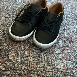 Svarta sneakers i skinn med vit sula - Snygga svarta sneakers från Lacoste i äkta skinn med vit platt sula och svarta snören. Skorna har en stilren design med perforerade detaljer baktill och bruna innersidor. Perfekta för dig som gillar en clean och klassisk look.