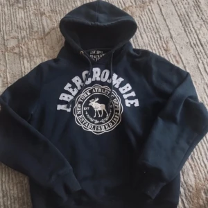 Mörkblå hoodie från Abercrombie - Snygg mörkblå hoodie från Abercrombie & Fitch med vit logga och tryck framtill. Tröjan har huva med snörning och är tillverkad i mjuk bomullsblandning (80% bomull, 20% polyester). Perfekt för chill dagar eller när du vill ha en sportig look.