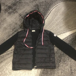 Svart dunväst med huva från Moncler - Svart dunväst från Moncler med huva och dragkedja. Västen har röda och vita detaljer längs dragkedjan och huvan, samt Moncler-logga på axeln. Tillverkad i polyester och dun, perfekt för lager-på-lager-stil.