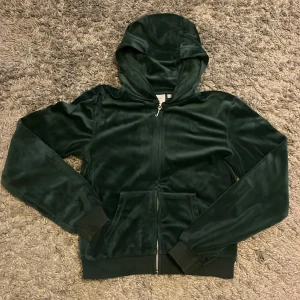 Mörkgrön velour hoodie Juicy Couture - Mörkgrön hoodie från Juicy Couture i mjuk velour med dragkedja och huva. Klassisk design med ribbade muddar, fickor fram och snygg metall-dragkedja. Perfekt för dig som vill ha en chill och trendig look. Byxorna i stl 15/16 och går tidigare varit upplagda med tejp upplägg men inget som syns utanpå  och tröjan stl 14/15. Tröjan har även någon fläck på fickan vet inte om de går bort. Obs kan även köpas separat! Om man köper separat kostar de 350 kr st 