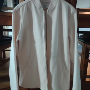 Vit bomberjacka från Ciszere XL - Snygg vit bomberjacka från Ciszere i slim fit. Jackan har dragkedja framtill, två sneda fickor och är tillverkad i en mix av akryl, polyester och ull. Perfekt för dig som gillar stilren och minimalistisk look.