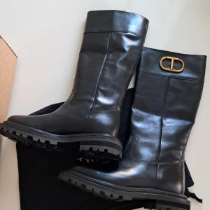 Svarta skinnstövlar Twinset med guldspänne - Snygga svarta stövlar från Twinset i äkta skinn med chunky sula och rund tå. Högt skaft med ikoniskt guldspänne upptill som ger en lyxig touch. Perfekta till höst- och vinterlooken, både stilrena och trendiga.