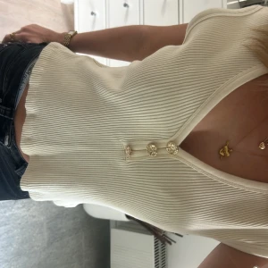 Ribbad vit topp med guldfärgade knappar - Säljer en ribbad vit topp från H&M i storlek M. Toppen har v-ringning och tre dekorativa guldfärgade knappar framtill. Modellen är croppad och ärmlös, perfekt för en chill och snygg look. Materialet är mjukt och stretchigt, troligtvis bomull eller syntet.