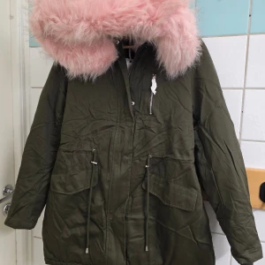 Olivgrön parkas med fluffig rosa foder - Parkasjacka i olivgrönt med stor huva och fluffigt rosa foder samt detaljer i vitt och svart. Jackan har snörning i midjan, dragkedja och flera fickor framtill. Insidan är mjuk och varm med pälsimitation i rosa, perfekt för kalla dagar.