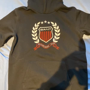 Svart hoodie från GANT med emblem - Svart hoodie från GANT med stort broderat emblem på bröstet i vitt och rött. Klassisk huva, magficka och långa ärmar. Perfekt för dig som gillar sportig och stilren look.