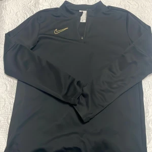 Svart Nike Dri-Fit långärmad tröja XL - Svart långärmad tröja från Nike i Dri-Fit material. Tröjan har en gul Nike-logga på bröstet och en halv dragkedja framtill. Perfekt för träning eller chill dagar. Snygg och sportig look med normal passform.
