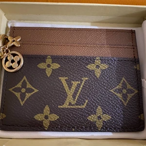 Louis Vuitton brun plånbok med monogram - Snygg plånbok från Louis Vuitton i brunt skinn med klassiskt monogrammönster och guldfärgad charm. Plånboken har flera kortfack och en lyxig känsla med ikoniska LV-detaljer. Perfekt för dig som vill ha något stilrent och exklusivt i fickan.