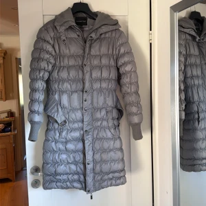 Grå lång pufferjacka Ilse Jacobsen - Snygg lång pufferjacka från Ilse Jacobsen i grått med quiltad design och metallicspänne i midjan. Jackan har hög mysig krage, ribbade muddar och både dragkedja och knappar framtill. Perfekt för kalla dagar med sin stilrena look och sköna passform.
