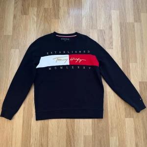  sweatshirt Tommy Hilfiger - Svart sweatshirt från Tommy Hilfiger med klassisk logga och text i guld över bröstet. Tröjan har en bred rand i vitt och rött framtill, rund hals och långa ärmar. Tillverkad i mjuk bomull för en skön och avslappnad stil.