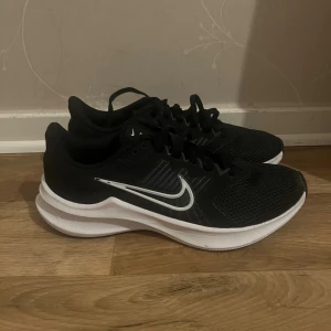 Svarta Nike sneakers - Snygga svarta Nike sneakers med en klassisk vit swoosh på sidan. Skorna har mesh-överdel för bra ventilation och snörning för perfekt passform. Perfekta för dig som gillar sportig stil och vill ha bekväma skor till träning eller vardag.