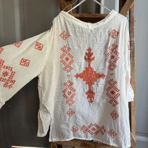 Vit bohemisk skjorta med broderi - Unik vit öppen tunika / topp från franska Charlie Joe i luftig bomull med orange broderade mönster på både fram- och baksida samt vida ärmar. Skjortan har en loose passform och långa ärmar med lätt utsvängda avslut. Perfekt för dig som gillar boho-vibe och detaljerad design.  Storlek S men passar XS - M 