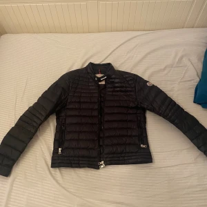 Svart Moncler dunjacka herr - Säljer en svart dunjacka från Moncler med klassisk quiltad design och diskret logga på ärmen. Jackan har dragkedja framtill, två sidofickor med dragkedja och en stilren, smal passform. Perfekt för dig som vill ha en lätt och snygg jacka till höst och vinter.