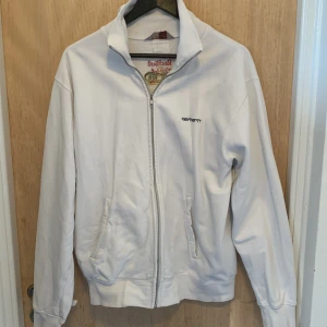 Vit ziptröja Carhartt x Red Bull Traffic Jam - Vit långärmad ziptröja från Carhartt med broderad logga på bröstet och Red Bull Traffic Jam-brodyr på ryggen. Tröjan har hög krage, två fickor fram och dragkedja hela vägen. Tillverkad i mjuk bomull, perfekt för chill dagar.