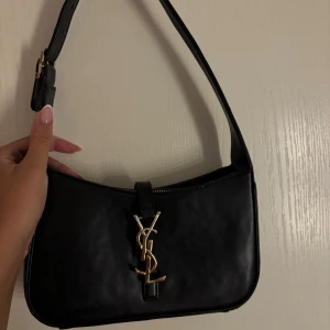 Ej äkta Svart axelväska från Saint Laurent - Snygg svart axelväska från Saint Laurent med ikoniskt YSL-emblem i guld på framsidan. Väskan är tillverkad i slätt skinn och har justerbart axelband med guldfärgat spänne där det står Saint Laurent Paris. Perfekt storlek för det viktigaste.