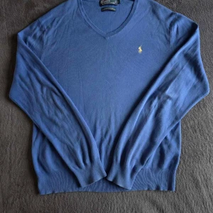 Blå v-ringad tröja från Polo Ralph Lauren - Snygg blå v-ringad tröja från Polo Ralph Lauren i 100% pima bomull. Tröjan har lång ärm, ribbade muddar och den klassiska broderade loggan i gult på bröstet. Perfekt för dig som gillar stilrena och tidlösa plagg.