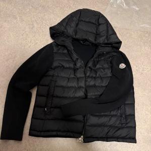 Moncler cardigan  - Svart pufferjacka från Moncler med quiltad front, stickade ärmar och huva. Jackan har dragkedja framtill, två fickor med dragkedja och Moncler-logga på ena ärmen. Perfekt för kyliga dagar och riktigt snygg streetstil. Skick 8/10, för mer bilder kom privat.