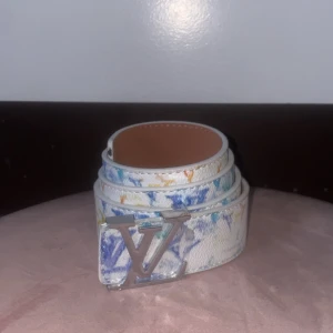 Fint färgat bälte - Säljer ett vitt bälte med färgstänk i blått, gult och orange. Bältet har en stor silverfärgad logga som spänne och är gjort i skinn. Perfekt för dig som vill sticka ut med en unik accessoar.