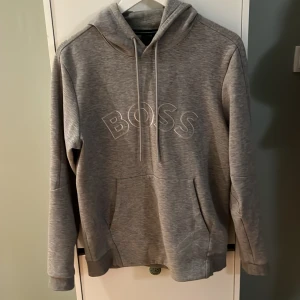 Grå hoodie från BOSS med broderad logga - Snygg grå hoodie från BOSS med broderad BOSS-logga på bröstet. Tröjan har huva med snörning, känguruficka och långa ärmar. Perfekt för en chill och stilren look.
