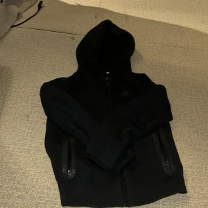 Svart nike tech fleece med dragkedja - Svart hoodie från Nike tech med hel dragkedja framtill och två fickor med dragkedja. Klassisk huva och diskret Nike-logga på bröstet. Perfekt för en avslappnad och sportig stil.