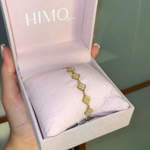 Guldarmband från HIMO med fyrklöver - Ett armband från HIMO, i guldton. Det ena sidan har glittrande fyrklöverformade länkar med små stenar, den andra sidan har svarta fyrklöver i guldinfattning. Perfekt accessoar för att lyfta din outfit med en lyxig touch.