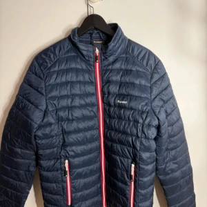 Mörkblå pufferjacka från Tuxer M - Snygg mörkblå pufferjacka från Tuxer med röda detaljer på dragkedjorna. Jackan har quiltad design, hög krage och två sidofickor med dragkedja. Perfekt för kyliga dagar och har en modern, sportig look.