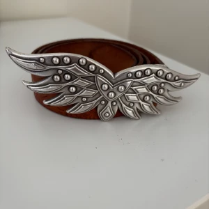 Brunt skärp med vingar från Saddler - Säljer ett brunt bälte i skinn från Saddler med en stor, detaljerad silverspänne i form av vingar. Bältet har präglat mönster längs hela remmen som ger en cool westernkänsla. Perfekt accessoar för att lyfta din outfit med en unik touch.