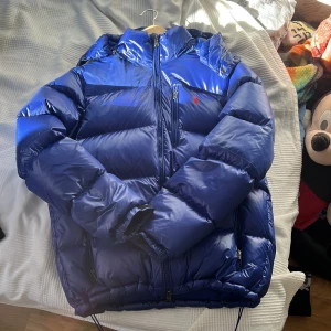 Blå pufferjacka från Polo Ralph Lauren - Snygg blå pufferjacka från Polo Ralph Lauren med röd broderad logga på bröstet. Jackan har huva, flera dragkedjefickor och justerbar nederkant med snörning. Perfekt för kalla dagar och riktigt skön att bära. Klassisk och sportig look. Lite för stor för mig då den är i storlek Large. Och jag är runt 182cm. 